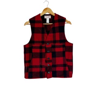 Puede incluir: Un chaleco de lana a cuadros rojos y negros con cuello en V y cierre de botones. El chaleco tiene dos bolsillos delanteros y est&aacute; colgado de una percha de madera. El patr&oacute;n es un cl&aacute;sico cuadro de b&uacute;falo.