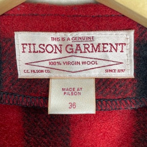 Op de afbeelding: Een rood en zwart geruit Filson kledinglabel met de tekst "This is a genuine Filson Garment 100% Virgin Wool C.C. Filson Co. Since 1897" en een kleiner wit label met de tekst "Made at Filson 36".