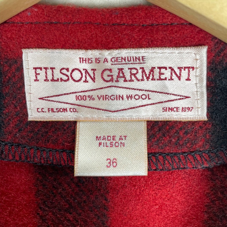 Op de afbeelding: Een rood en zwart geruit Filson kledinglabel met de tekst "This is a genuine Filson Garment 100% Virgin Wool C.C. Filson Co. Since 1897" en een kleiner wit label met de tekst "Made at Filson 36".