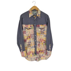 May include: A long-sleeved button-down shirt with a patchwork design. The shirt features a denim-like blue fabric on the sleeves and upper chest, and a colorful patchwork pattern on the front. The patchwork includes squares of red, yellow, and purple.
