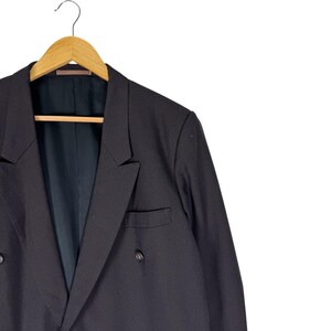Op de afbeelding: Een zwarte blazer met dubbele rij knopen, hangend aan een houten kleerhanger. De blazer heeft twee rijen knopen en een gekerfde revers. De binnenvoering is donker van kleur.