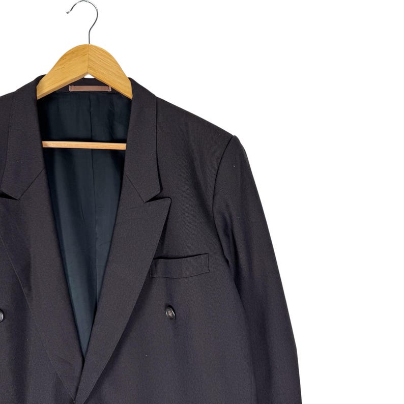 Op de afbeelding: Een zwarte blazer met dubbele rij knopen, hangend aan een houten kleerhanger. De blazer heeft twee rijen knopen en een gekerfde revers. De binnenvoering is donker van kleur.