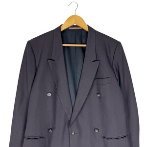 Op de afbeelding: Een zwarte double-breasted blazer hangt aan een houten hanger. De blazer heeft een klassieke revers, twee rijen knopen en twee voorzakken. De binnenvoering is donker van kleur.