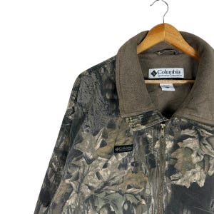 May include: A camouflage Columbia Sportswear Company jacket with a brown fleece collar and a full-length zip. The jacket features a leaf pattern in shades of green, brown, and grey. The Columbia logo is visible on the collar and the chest.