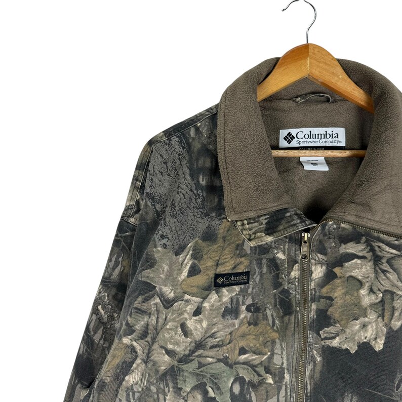 May include: A camouflage Columbia Sportswear Company jacket with a brown fleece collar and a full-length zip. The jacket features a leaf pattern in shades of green, brown, and grey. The Columbia logo is visible on the collar and the chest.