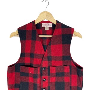 Op de afbeelding: Rood en zwart geruit flanellen vest met twee grote zakken en een knoopsluiting. Het vest heeft een Filson Garment-label.