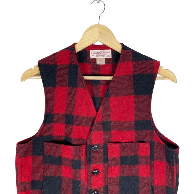 Op de afbeelding: Rood en zwart geruit flanellen vest met twee grote zakken en een knoopsluiting. Het vest heeft een Filson Garment-label.