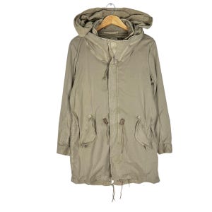 May include: A khaki-colored hooded parka jacket with a drawstring waist and pockets. The jacket is hanging on a wooden hanger against a white background. The parka has a long length and a relaxed fit.