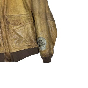 May include: A vintage brown leather bomber jacket with two front pockets and a brown ribbed hem and cuffs. A circular patch with an illustration is on the sleeve. The jacket has a distressed appearance.