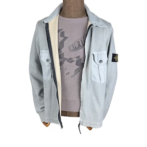 Könnte beinhalten: Hellblaue Cordjacke mit durchgehendem Reißverschluss, zwei Vordertaschen und einem kleinen Logoaufnäher am linken Ärmel. Die Jacke wird über einem grau gemusterten Pullover getragen. Die Jacke hat ein helles Futter.