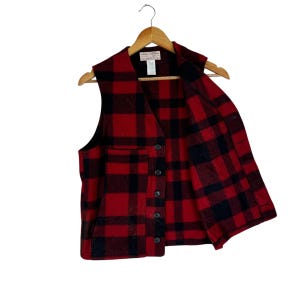 Puede incluir: Chaleco a cuadros rojos y negros con cierre de botones, colgado de una percha de madera. El chaleco tiene un bolsillo en el lado izquierdo y una etiqueta en el interior. Hecho de un material grueso y c&aacute;lido.