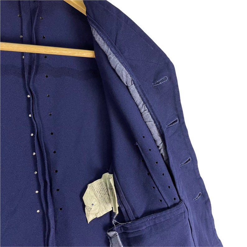 Puede incluir: Una chaqueta azul marino con cierre de botones y un solo bolsillo en el lado izquierdo. La chaqueta tiene un interior forrado y una etiqueta con texto japon&eacute;s.