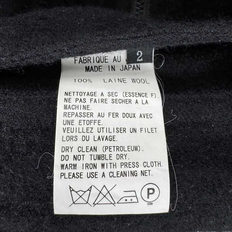 K&ouml;nnte beinhalten: Ein wei&szlig;es Pflegeetikett mit schwarzem Text und Symbolen. Das Etikett lautet "Fabrique au 2 Made in Japan 100% Laine Wool" und enth&auml;lt Anweisungen f&uuml;r die chemische Reinigung, das B&uuml;geln und das Waschen. Das Etikett enth&auml;lt auch ein "P"-Symbol.