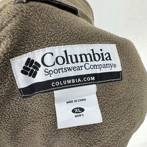 May include: Close-up of a brown fleece jacket with the Columbia Sportswear Company label. The label is white with black text and a logo. A smaller tag indicates size XL and "Men's". The jacket is made in China.