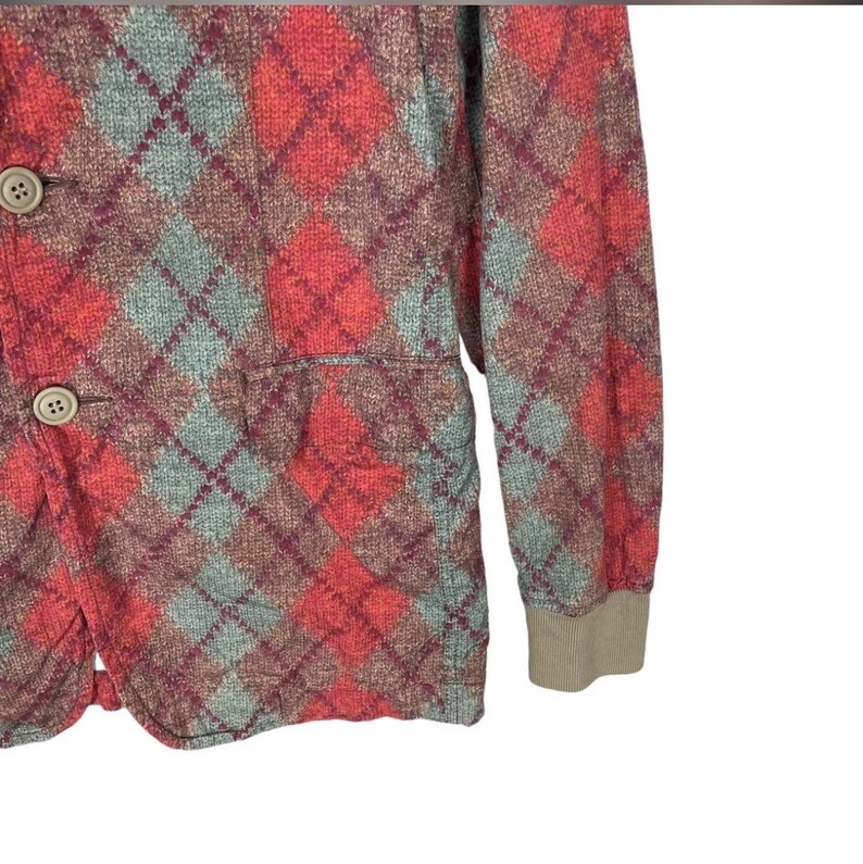 May include: A red, teal, and brown argyle patterned jacket with two buttons and a pocket. The jacket has a beige ribbed cuff on the sleeve. The argyle pattern is a classic design.