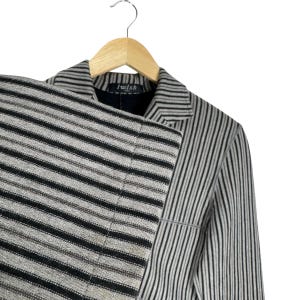Op de afbeelding: Een gestreepte blazer en bijpassende broek in zwart, grijs en beige, hangend aan een houten hanger. De jas heeft verticale strepen, terwijl de broek horizontale strepen heeft. Het label in de jas luidt "I wish".