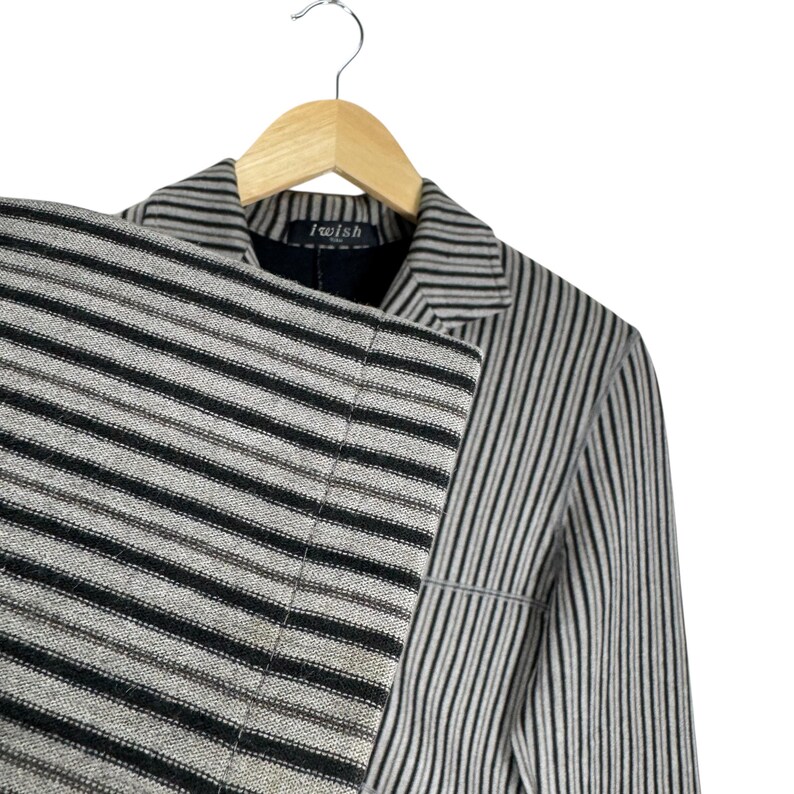 Op de afbeelding: Een gestreepte blazer en bijpassende broek in zwart, grijs en beige, hangend aan een houten hanger. De jas heeft verticale strepen, terwijl de broek horizontale strepen heeft. Het label in de jas luidt "I wish".