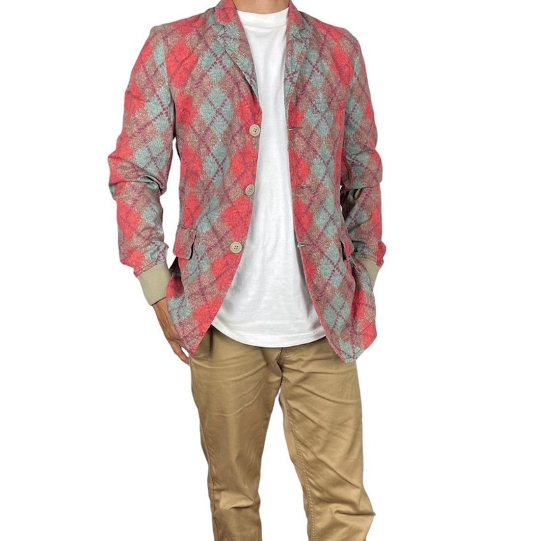 May include: A red, grey, and blue argyle patterned blazer with three beige buttons. The blazer has two front pockets and a beige cuff. The blazer is worn over a white t-shirt and paired with khaki trousers.