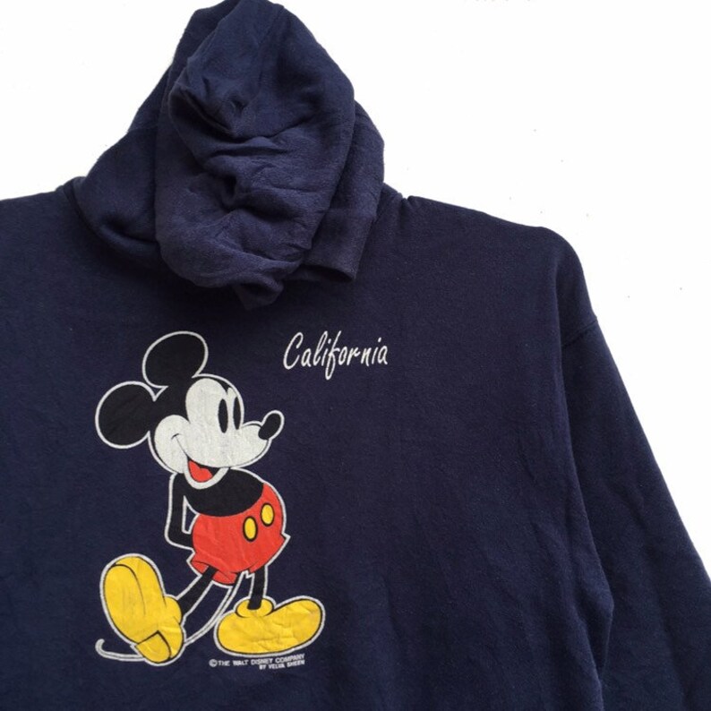 K&ouml;nnte beinhalten: Ein marineblauer Kapuzenpullover mit einem Cartoon-Bild von Mickey Mouse und dem Wort "California" auf der Vorderseite. Die Copyright-Informationen von The Walt Disney Company sind unten im Bild aufgedruckt.