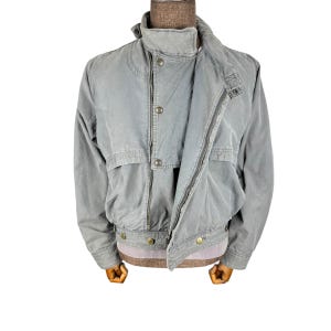 May include: A light grey bomber jacket with a zip-up front, button details, and a ribbed hem. The jacket has a stand-up collar and two front pockets. The jacket is displayed on a mannequin with wooden arms.
