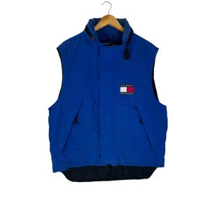May include: A blue sleeveless puffer vest with a black zipper and a Tommy Hilfiger logo patch on the front. The logo is a red, white, and blue flag with the text "Tommy Hilfiger" underneath.