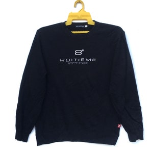 以下が含まれることがあります： 白いロゴ「8° Huitième Sports Studio」と袖に赤いタグが付いた黒い長袖スウェットシャツです。
