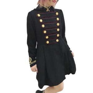 Può includere: Cappotto nero in stile militare con bottoni e finiture dorate. Il cappotto presenta un collo alto e una gonna svasata. Le maniche lunghe hanno ricami dorati vicino ai polsini. Il cappotto ha un design doppiopetto con sei bottoni dorati.