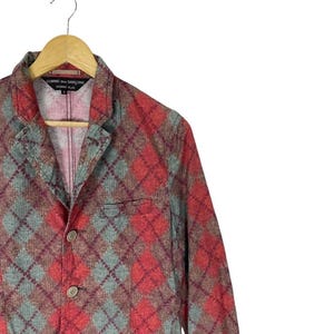 May include: A red, grey, and brown argyle patterned blazer with two buttons and a notched lapel. The blazer is hanging on a wooden hanger. The label reads "Comme des Garcons Homme Plus."