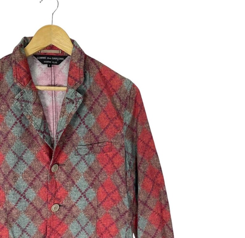 May include: A red, grey, and brown argyle patterned blazer with two buttons and a notched lapel. The blazer is hanging on a wooden hanger. The label reads "Comme des Garcons Homme Plus."