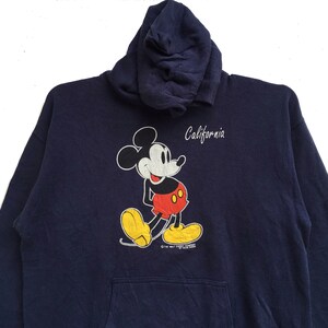 K&ouml;nnte beinhalten: Ein marineblauer Kapuzenpullover mit einem Mickey-Mouse-Motiv und dem Schriftzug "California" auf der Vorderseite.