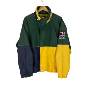 Puede incluir: Una chaqueta verde, azul marino y amarilla con cierre de cremallera. La chaqueta tiene un logotipo de Tommy Hilfiger Sailing Gear en la manga derecha.