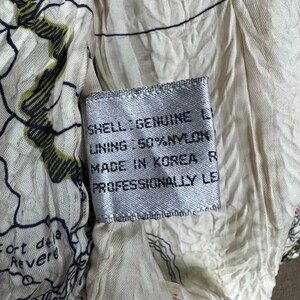 May include: Close-up of a tag with text on a cream-colored fabric. The tag reads "SHELL: GENUINE LI, LINING: 50% NYLON, MADE IN KOREA R, PROFESSIONALLY LE". The fabric has a map-like design with green and black accents.