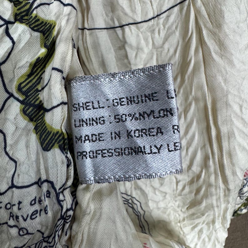 May include: Close-up of a tag with text on a cream-colored fabric. The tag reads "SHELL: GENUINE LI, LINING: 50% NYLON, MADE IN KOREA R, PROFESSIONALLY LE". The fabric has a map-like design with green and black accents.