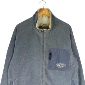 Puede incluir: Chaqueta polar gris con cremallera completa y cuello alto. La chaqueta tiene un bolsillo azul oscuro en el pecho con un peque&ntilde;o logotipo. El interior del cuello es beige. La chaqueta est&aacute; colgada de una percha de madera.