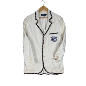 Peut inclure: Blazer crème avec bordure marine et dorée, orné d'un écusson brodé marine sur la poitrine. Le blazer a un design classique avec trois boutons et deux poches avant. Il est suspendu à un cintre en bois.