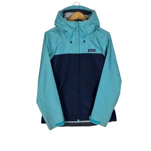 May include: A teal and navy blue hooded jacket with a zip closure. The jacket features a small logo patch on the chest and adjustable cuffs. The jacket is designed for outdoor activities and is made of a lightweight, water-resistant material.