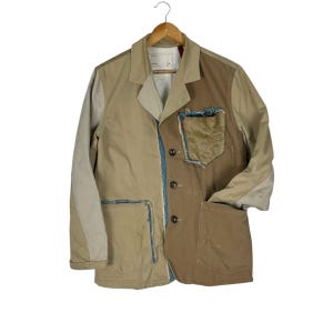 Puede incluir: Un blazer patchwork en tonos beige y marrón, con detalles de mezclilla y un bolsillo. La chaqueta tiene una botonadura y está colgada de una percha de madera. El diseño incorpora telas y texturas contrastantes.