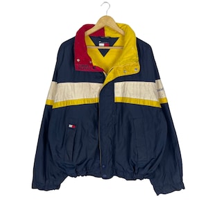 Puede incluir: Una chaqueta azul marino, amarilla y blanca a rayas con cuello rojo y la marca Tommy Hilfiger. La chaqueta tiene cierre de cremallera y un cordón en la parte inferior.