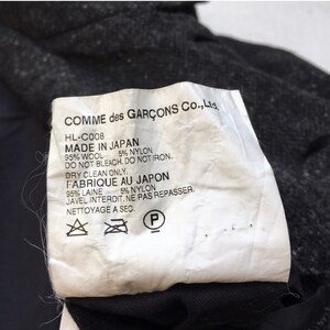 May include: White clothing label with black text that reads "COMME des GAR&Ccedil;ONS Co., Ltd. HL-C008 MADE IN JAPAN 95% WOOL 5% NYLON DO NOT BLEACH. DO NOT IRON. DRY CLEAN ONLY. FABRIQUE AU JAPON 95% LAINE 5% NYLON JAVEL INTERDIT. NE PAS REPASSER. NETTOYAGE A SEC." The label has three symbols: a triangle with an X inside, a circle with a line through it, and a circle with a P inside.