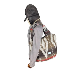 May include: A brown and white patterned tote bag with a red trim is slung over the shoulder. The bag features a geometric design. The person is wearing a brown aviator hat with ear flaps and a gray long-sleeved shirt.