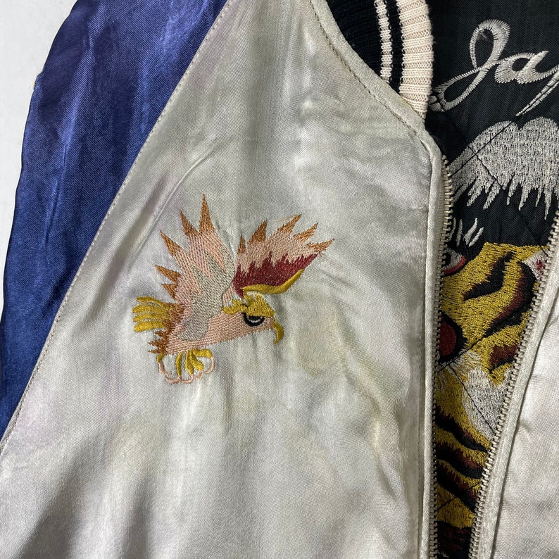 May include: A white satin bomber jacket with a blue satin sleeve and embroidered eagle on the front. The other side of the jacket features a tiger design and the word "Japan" in black.
