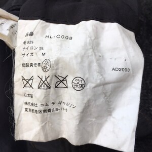 May include: A white tag with black text and symbols. The tag includes the text "HL-C008", "毛 95%", "ナイロン 5%", "サイズ M", "縫製責任者", "日本製", "株式会社 コムデギャルソン", "東京都港区南青山5-11-5", and "AD2003". The tag also includes symbols for washing instructions.