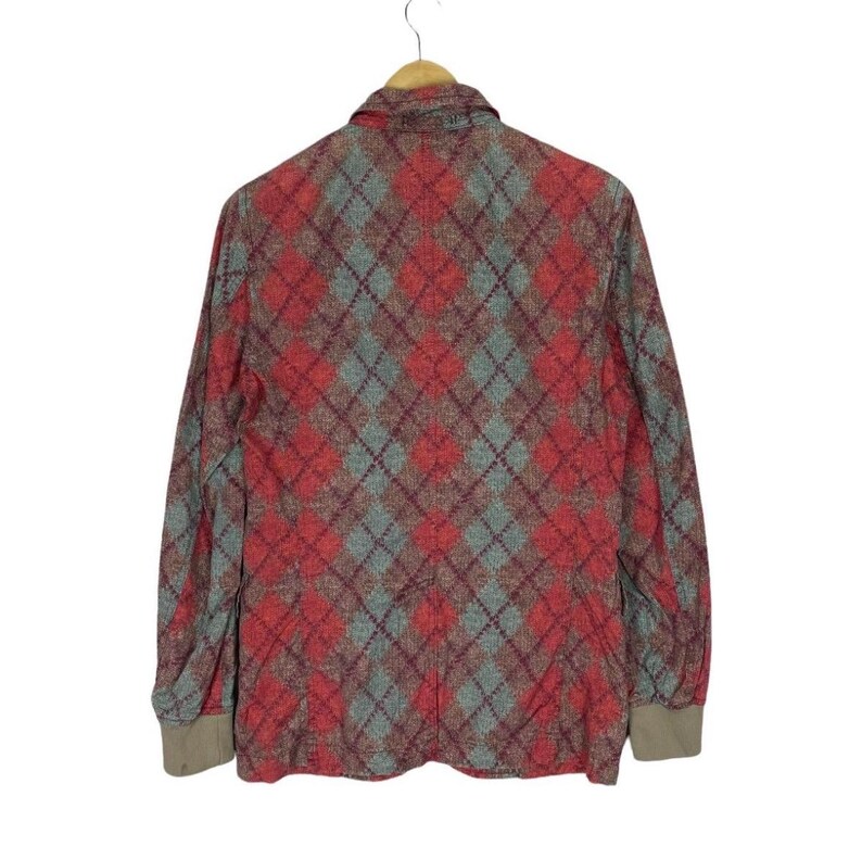 May include: A long-sleeved jacket with a red and grey argyle pattern. The jacket is suspended on a wooden hanger. The cuffs are a solid tan colour. The background is white.