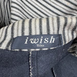 Op de afbeelding: Close-up van een zwart kledingstuk met een gestreepte voering. Het label luidt "i wish" in witte letters, met daaronder "Ys'bis". Het kledingstuk heeft een rits en lijkt gemaakt van een donkere, mogelijk marineblauwe stof.