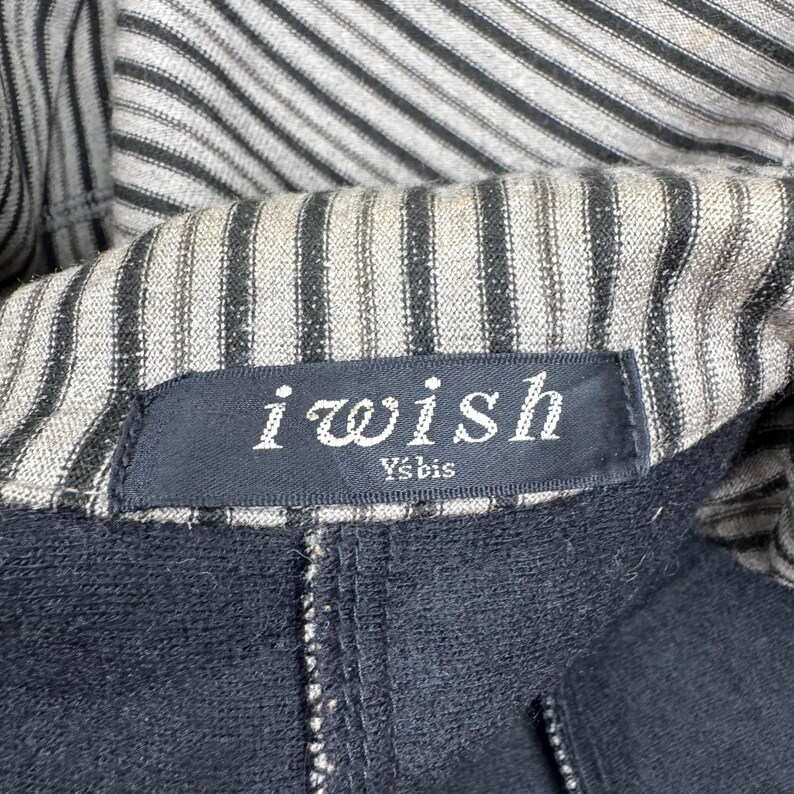 Op de afbeelding: Close-up van een zwart kledingstuk met een gestreepte voering. Het label luidt "i wish" in witte letters, met daaronder "Ys'bis". Het kledingstuk heeft een rits en lijkt gemaakt van een donkere, mogelijk marineblauwe stof.