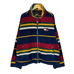 Puede incluir: Chaqueta polar a rayas con cremallera completa. La chaqueta presenta rayas horizontales en azul marino, burdeos, amarillo y blanco. El logotipo de Tommy Hilfiger está en el pecho.