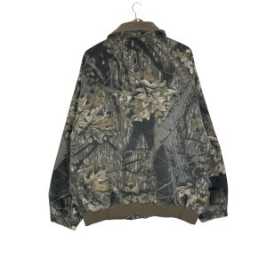 May include: A camouflage jacket with a brown collar and cuffs. The jacket features a pattern of green, brown, and grey, resembling a forest. The jacket is hanging on a metal hanger.