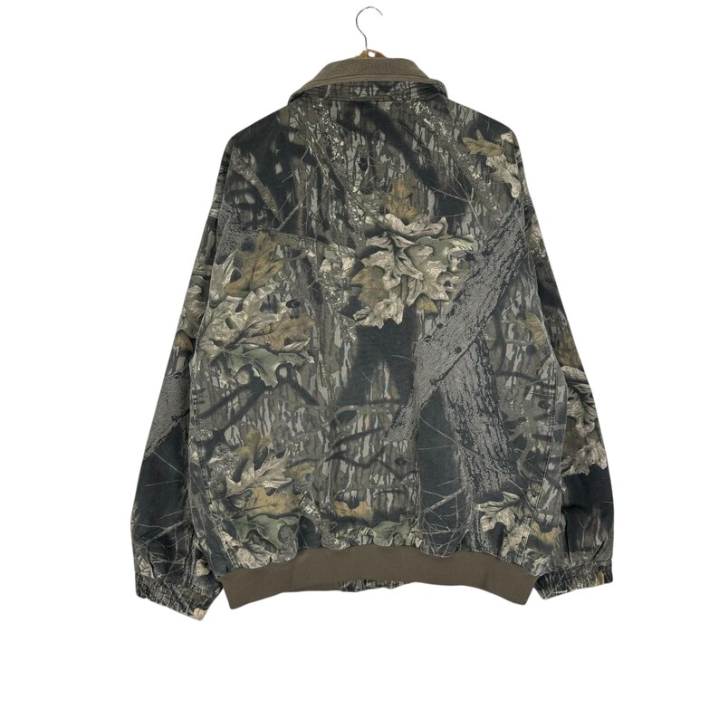 May include: A camouflage jacket with a brown collar and cuffs. The jacket features a pattern of green, brown, and grey, resembling a forest. The jacket is hanging on a metal hanger.