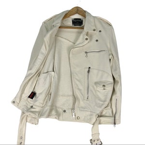 May include: Cream-colored, open motorcycle jacket with a soft, off-white interior lining. The jacket features multiple silver zippers, snap closures, and a belt. The brand name "Hysteric" is visible on a tag. The jacket is hanging on a wooden hanger.