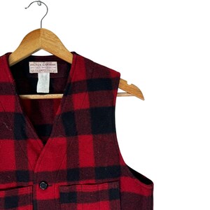 Puede incluir: Un chaleco a cuadros rojos y negros con cuello en V, colgado de una percha de madera. El chaleco tiene dos bolsillos delanteros y un cierre de bot&oacute;n. La etiqueta dice "FILSON GARMENT".
